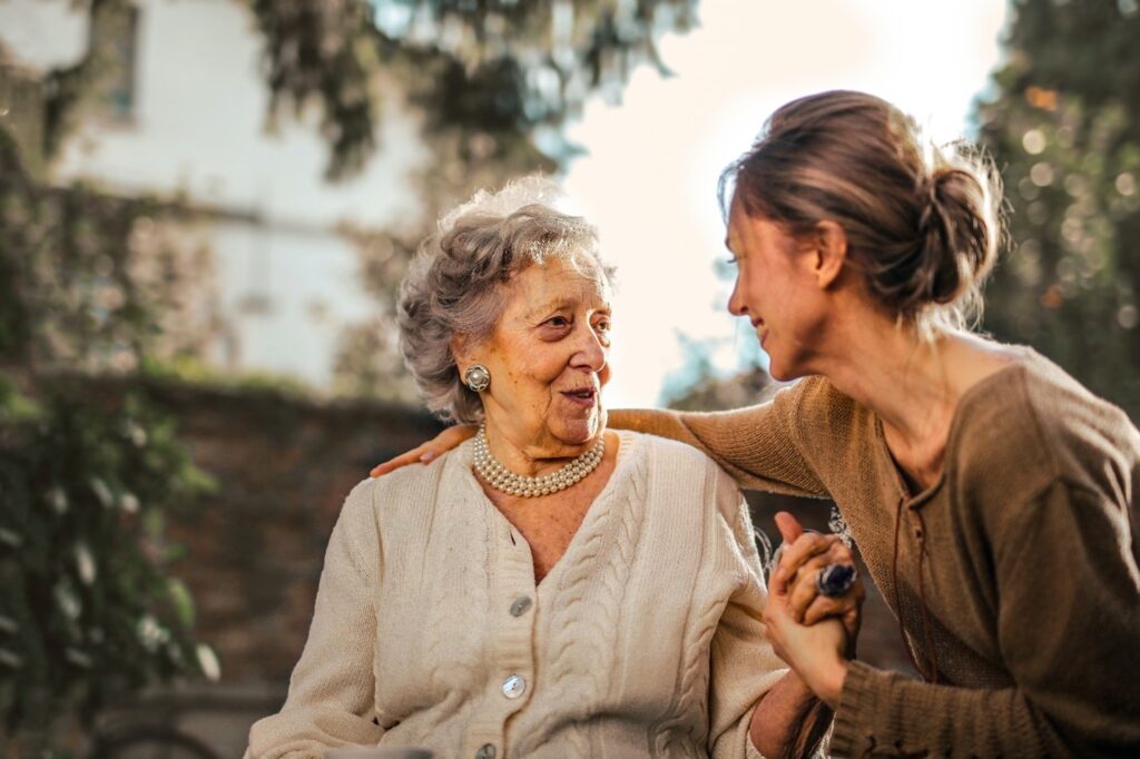 Image of two ladies embracing. The older lady is on the left wearing a white cardigan with a peral necklace. She is holding hands and smiling at a younger lady with brown hair who has her arm around her shoulder.