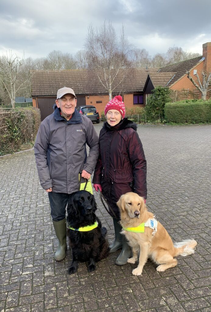 This article image is a picture of Roger and Margaret. They are standing side by side in front of a lovely brick house. It’s an overcast day and they are wearing coats and hats. They’re each holding onto their guide dogs who are sitting in front of them.